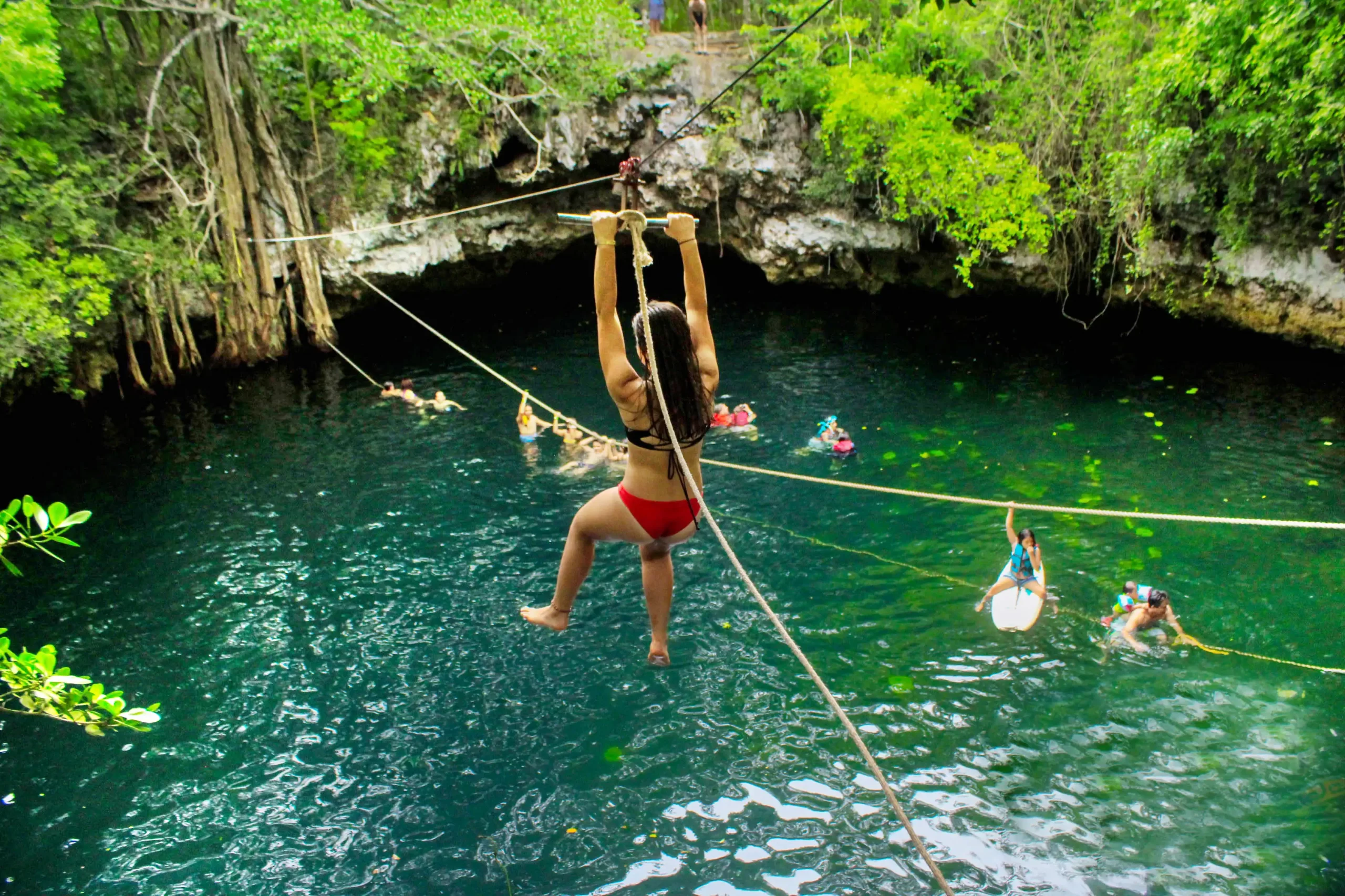 Woman ziplining over a cenote, holding onto the rope with both hands, with the vibrant green jungle and crystal-clear cenote water below.