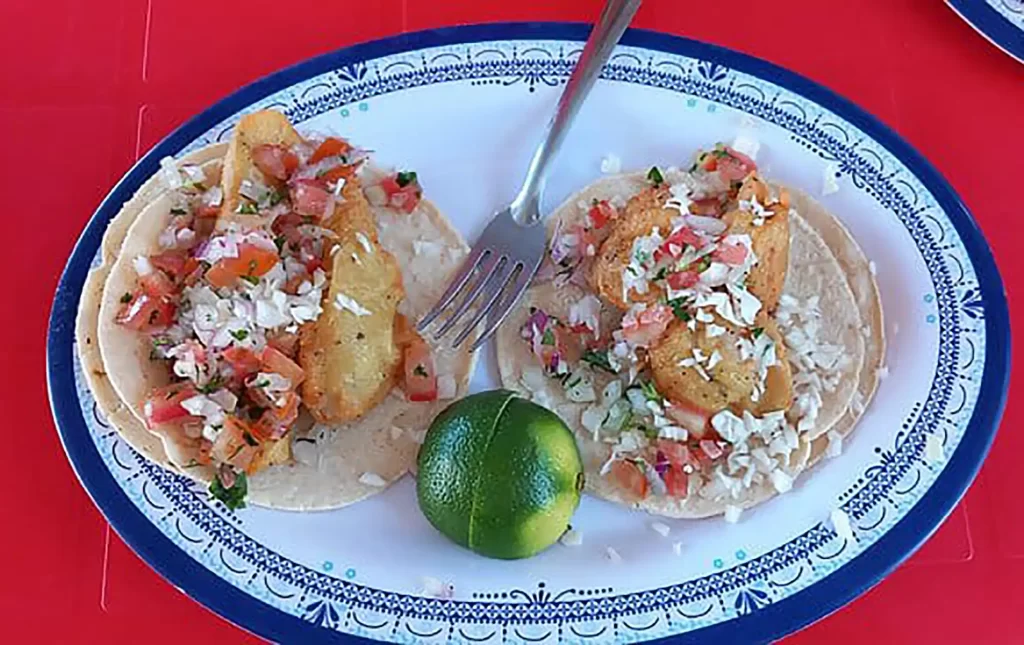 Two crispy fish tacos topped with fresh pico de gallo, served with a lime wedge.