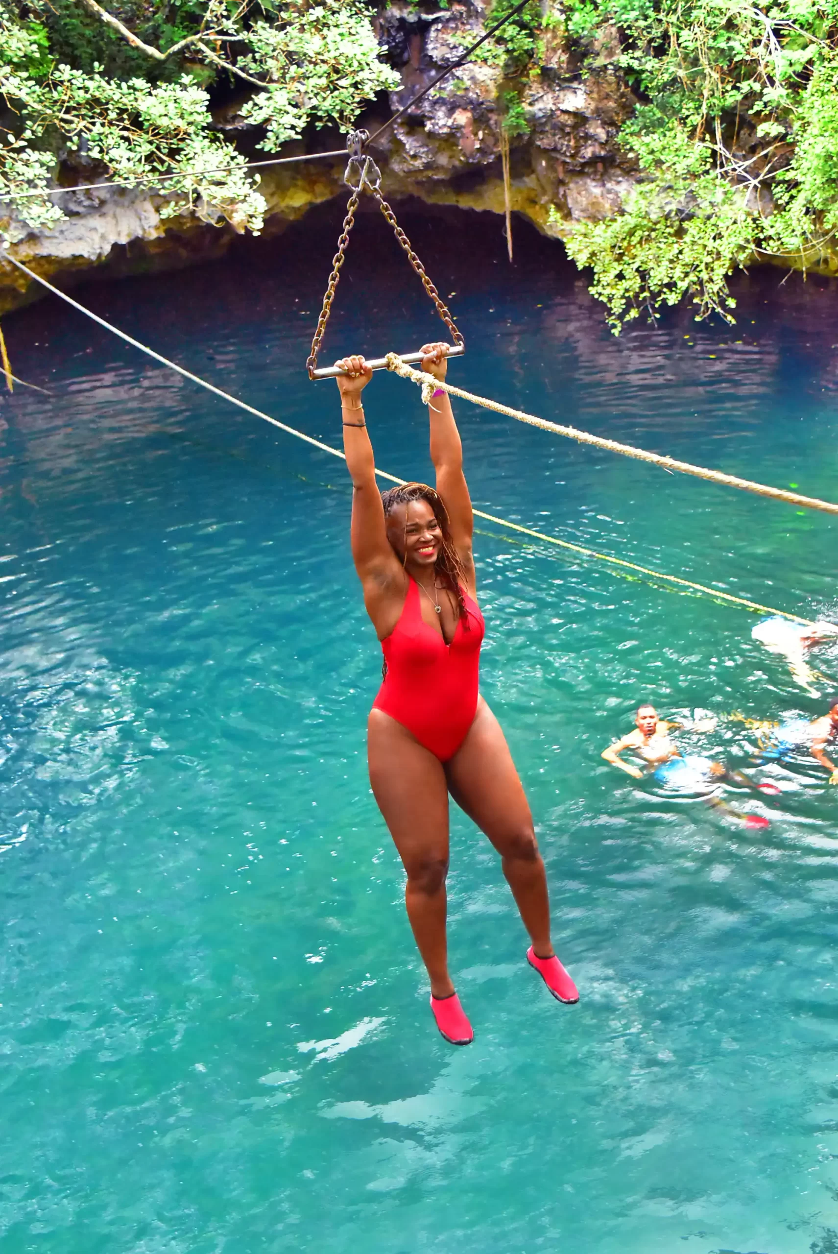 Woman in red swimsuit swinging on a rope over a crystal-clear cenote in Cancun.