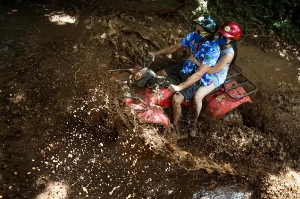 Two individuals riding an ATV through a muddy puddle, causing a large splash as they speed through the jungle terrain in Cancun.