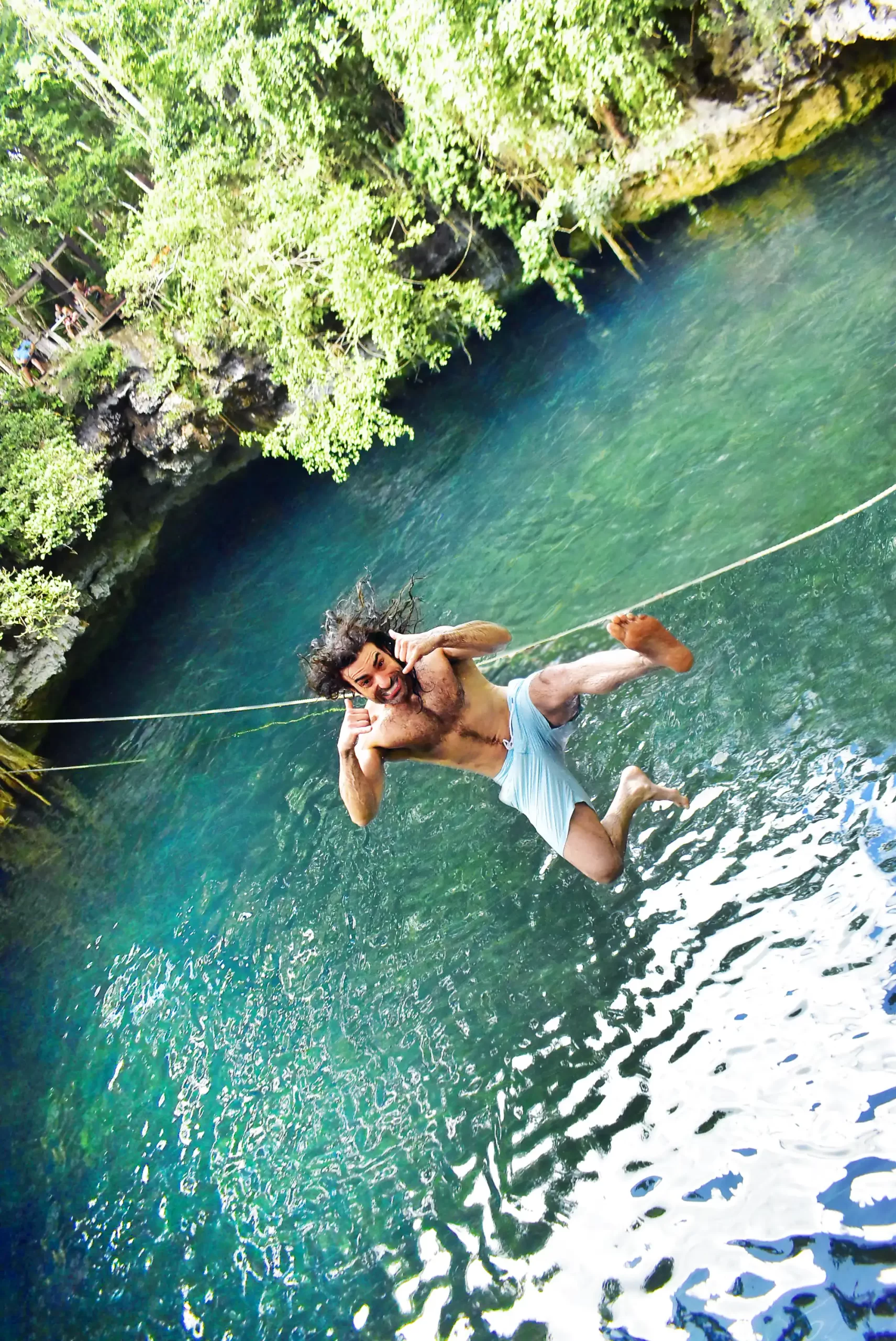 Man on a rope swing over a cenote with lush greenery surrounding the clear blue water.