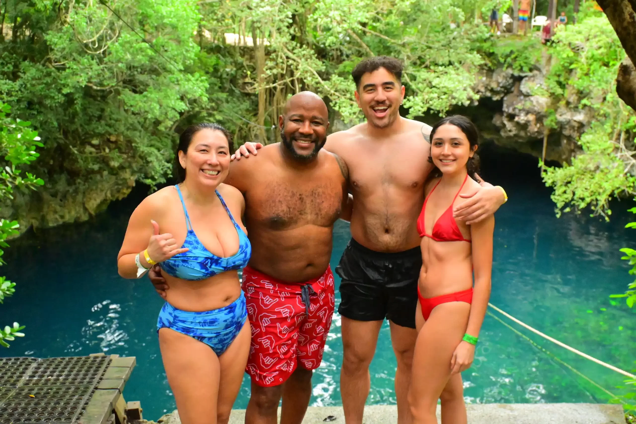 Group of friends posing by a cenote, smiling and wearing swimsuits with a backdrop of natural green and blue scenery