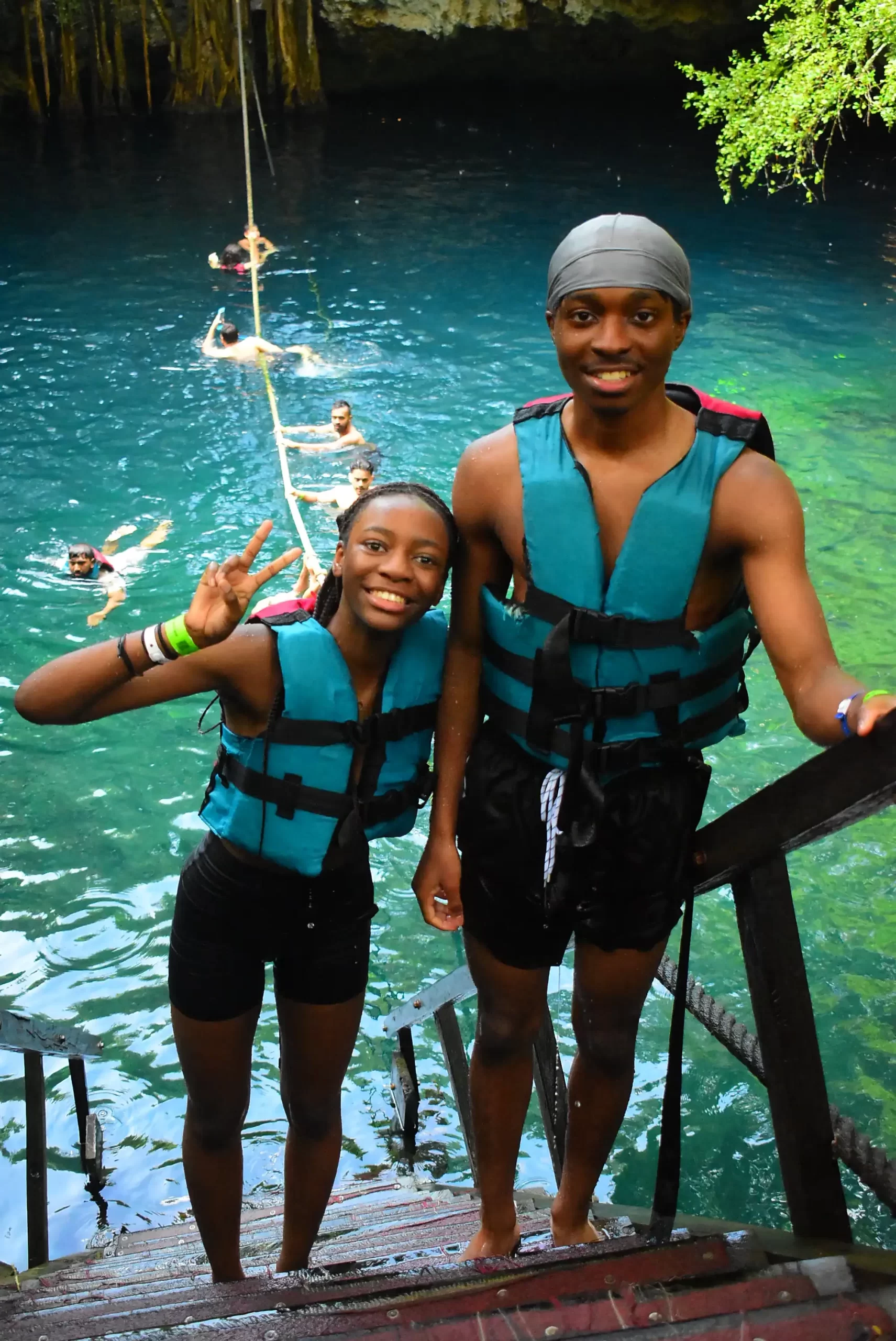Two friends in life jackets posing by a cenote with others swimming in the background in Cancun.