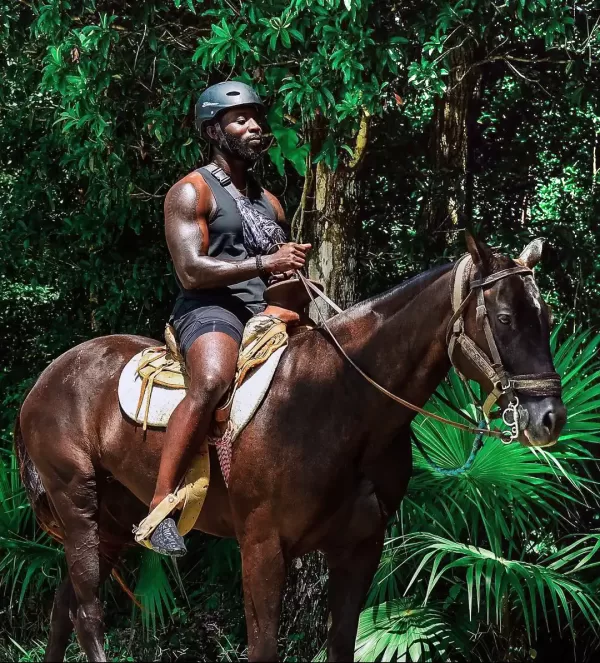 Man on horseback in a tropical forest, wearing a helmet and black tank top, confidently guiding the horse through the jungle