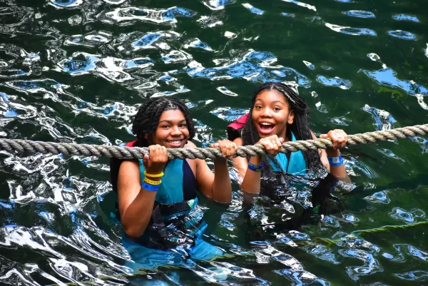Two girls hanging from a rope over water in a cenote, smiling and wearing life jackets.