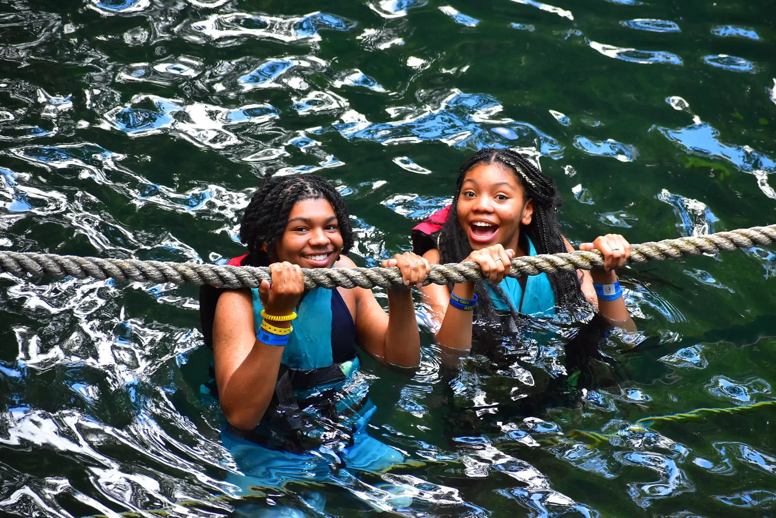 Two girls hanging from a rope over water in a cenote, smiling and wearing life jackets.