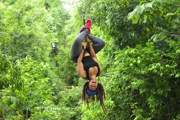 Woman hanging upside down on a zipline in the jungle, wearing a blue bandana, black top, and gray leggings, smiling as she enjoys the thrill.
