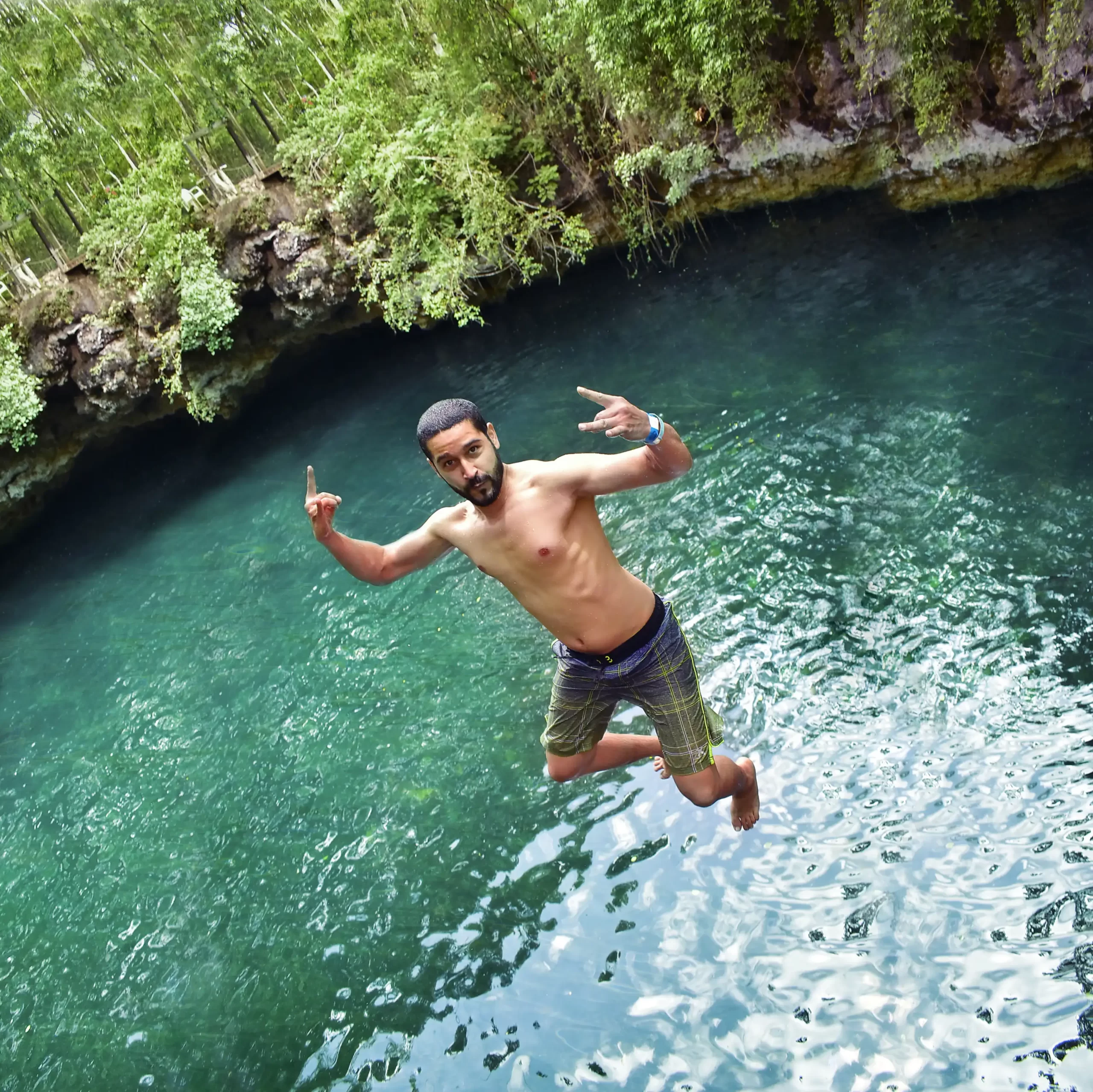 Man mid-jump into a cenote, making a peace sign with both hands, enjoying the water adventure