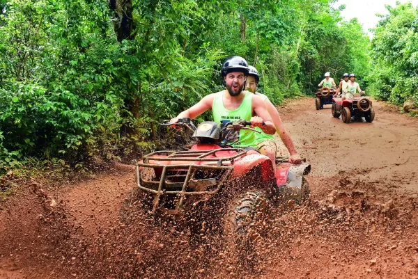 Man driving an ATV through a muddy jungle trail, splashing mud while leading a group of riders.