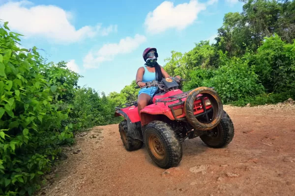Woman riding an ATV through a dirt trail surrounded by lush green foliage.