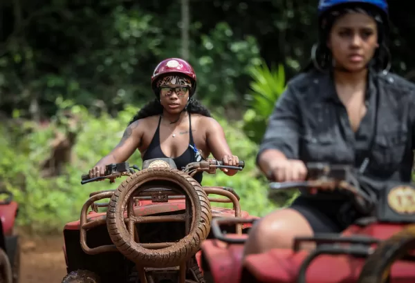 Two people riding ATVs through a forest, one wearing a red helmet and the other a blue helmet.