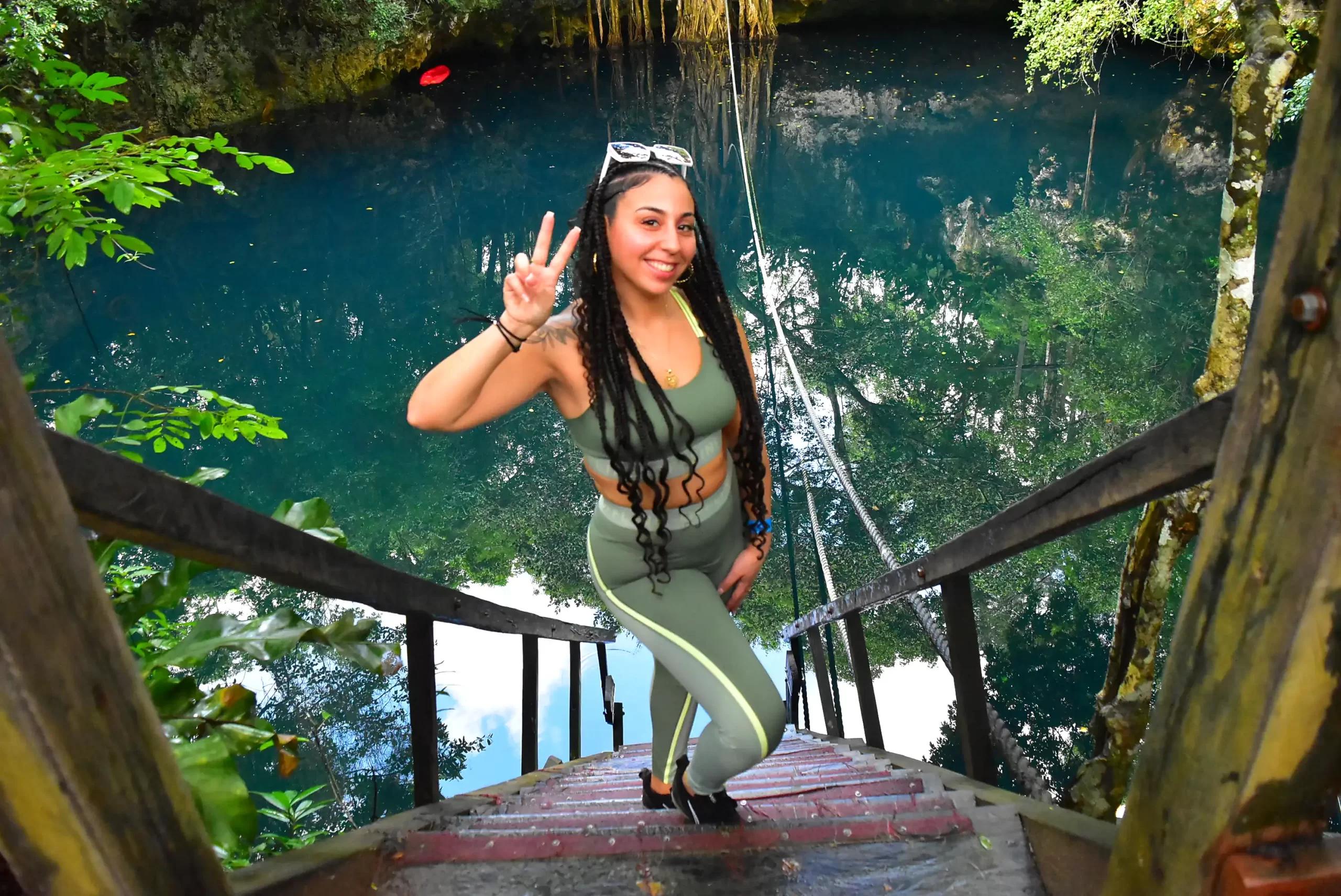 A woman in a green outfit standing on stairs, flashing a peace sign with a serene cenote in the background.