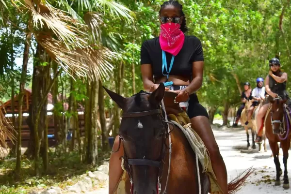 Person riding a horse with a pink bandana covering their face, wearing sunglasses and a black outfit in a tropical environment.