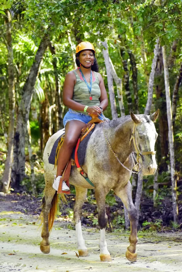 Smiling person in green camouflage top and helmet riding a light-colored horse along a forest trail.