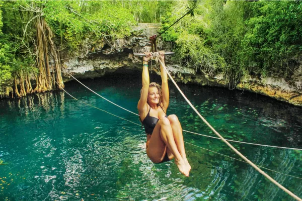 A woman zip-lining over a turquoise cenote in a tropical forest.