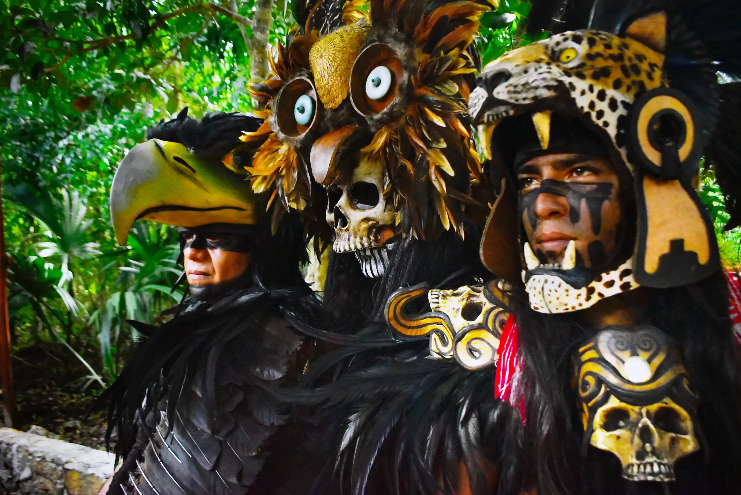 Three performers in animal-themed costumes, one in a bird mask, another in a skull and feathers, and a third in a jaguar mask.