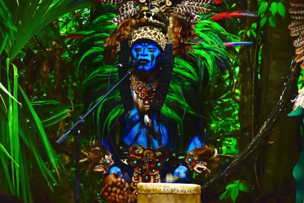 A performer painted blue and dressed in vibrant green and feathered costume playing a drum.