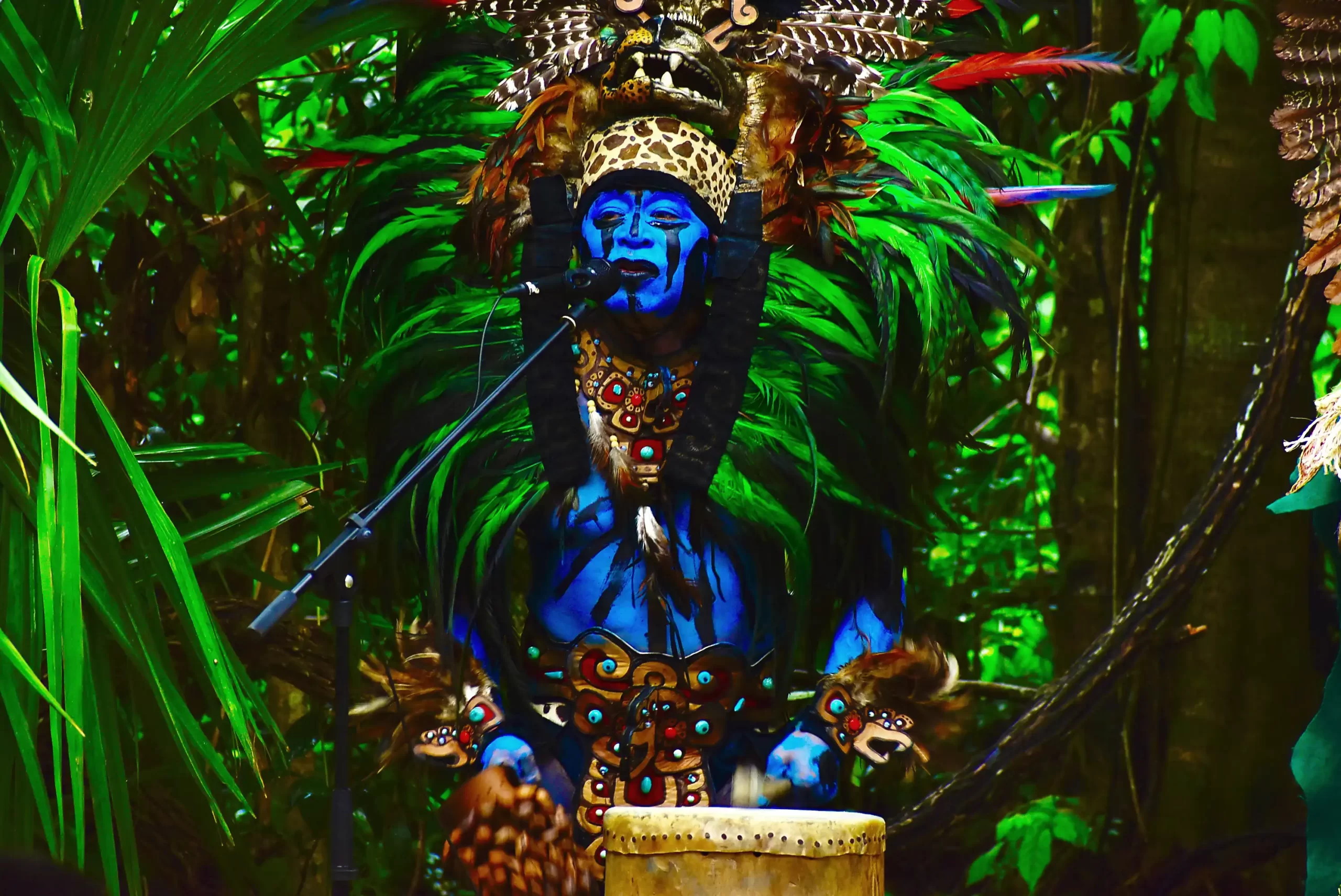 A performer painted blue and dressed in vibrant green and feathered costume playing a drum.