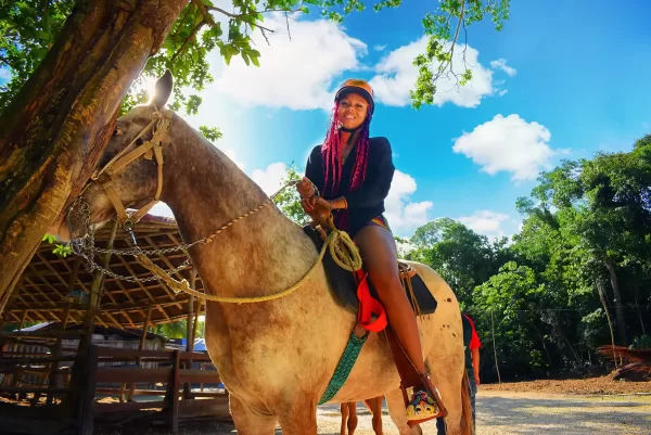 A woman with bright red braids, wearing an orange helmet, is riding a gray horse under a sunny sky. The horse is standing next to a tree, and the background features greenery and a blue sky with fluffy clouds.