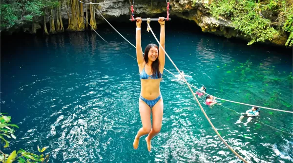 A woman in a blue bikini smiles as she hangs from a zipline above a clear blue cenote surrounded by lush green trees. In the background, other people are swimming in the cenote.