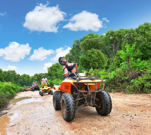 A woman on an ATV is having fun while riding through a muddy trail with a bright blue sky and lush greenery in the background.