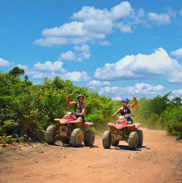 Two ATV riders showing excitement as they speed down a dirt trail surrounded by greenery and a sunny blue sky with fluffy white clouds.