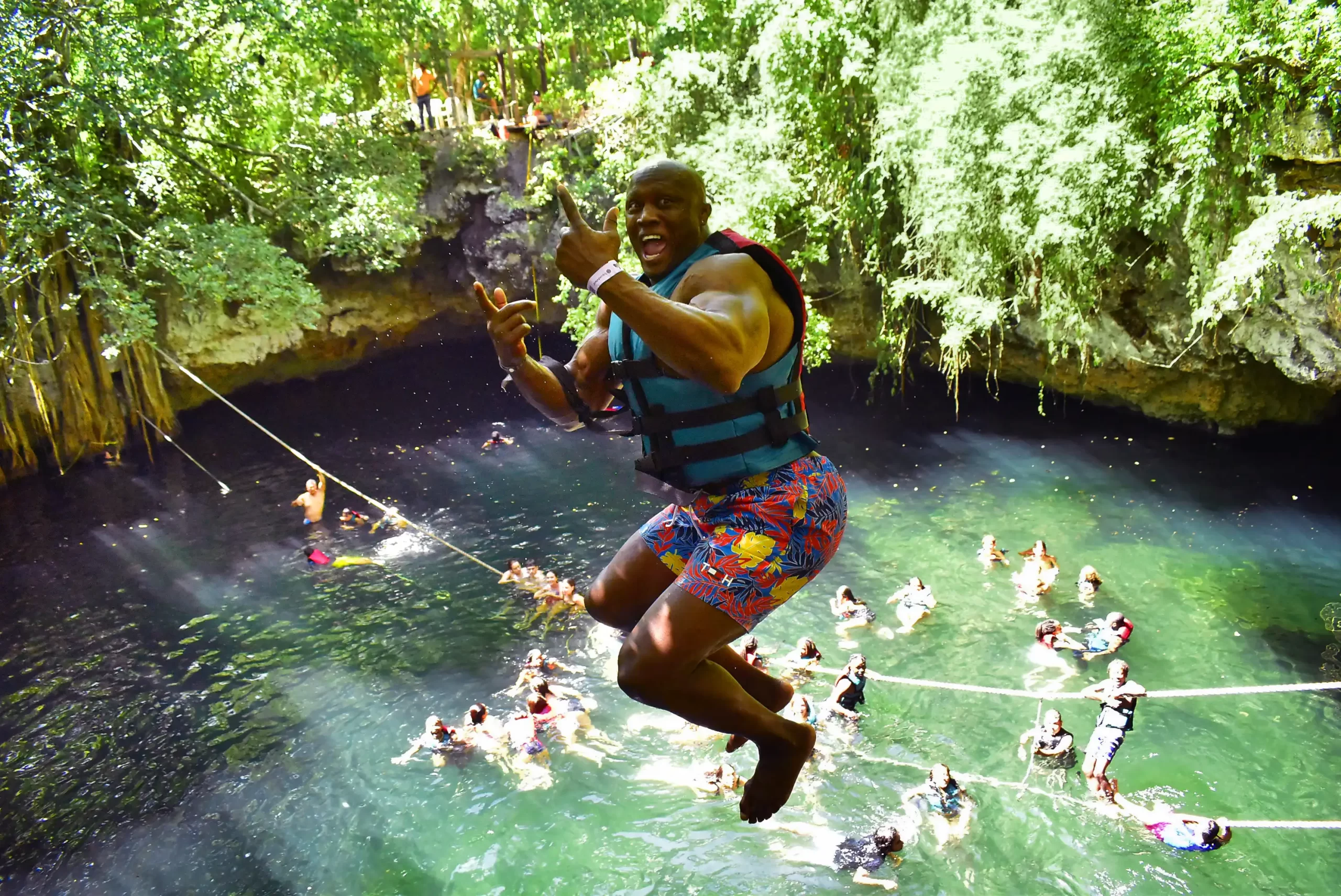 A man jumping into a cenote, giving a thumbs-up as he splashes into the crystal-clear water, with people swimming below.